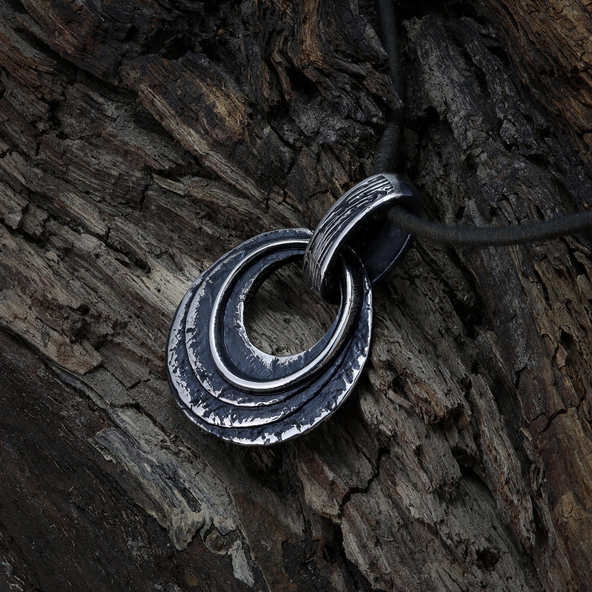 A textured, layered sterling silver pendant featuring two concentric circles, strung on a black leather cord, set against an ancient, weathered tree bark.