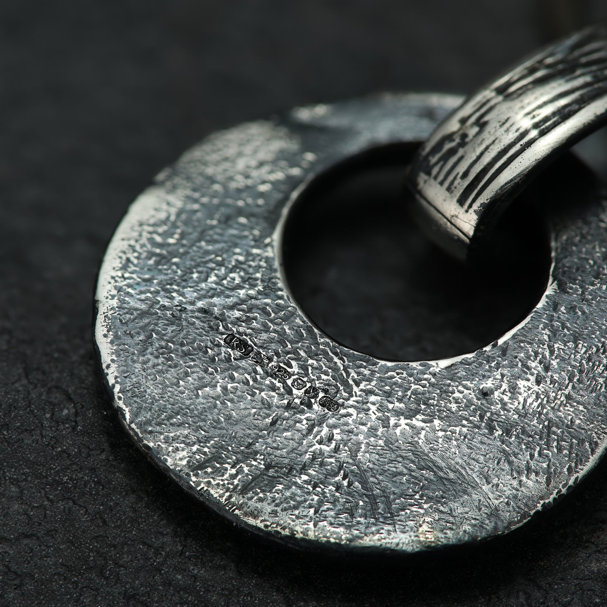 Close-up of a textured silver circular pendant with a hammered finish, highlighting the hallmark and the intricate craftsmanship, against a dark, matte background.